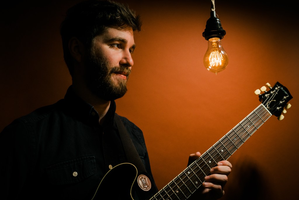 Max Light holding guitar and looking at an exposed lightbulb. Red background