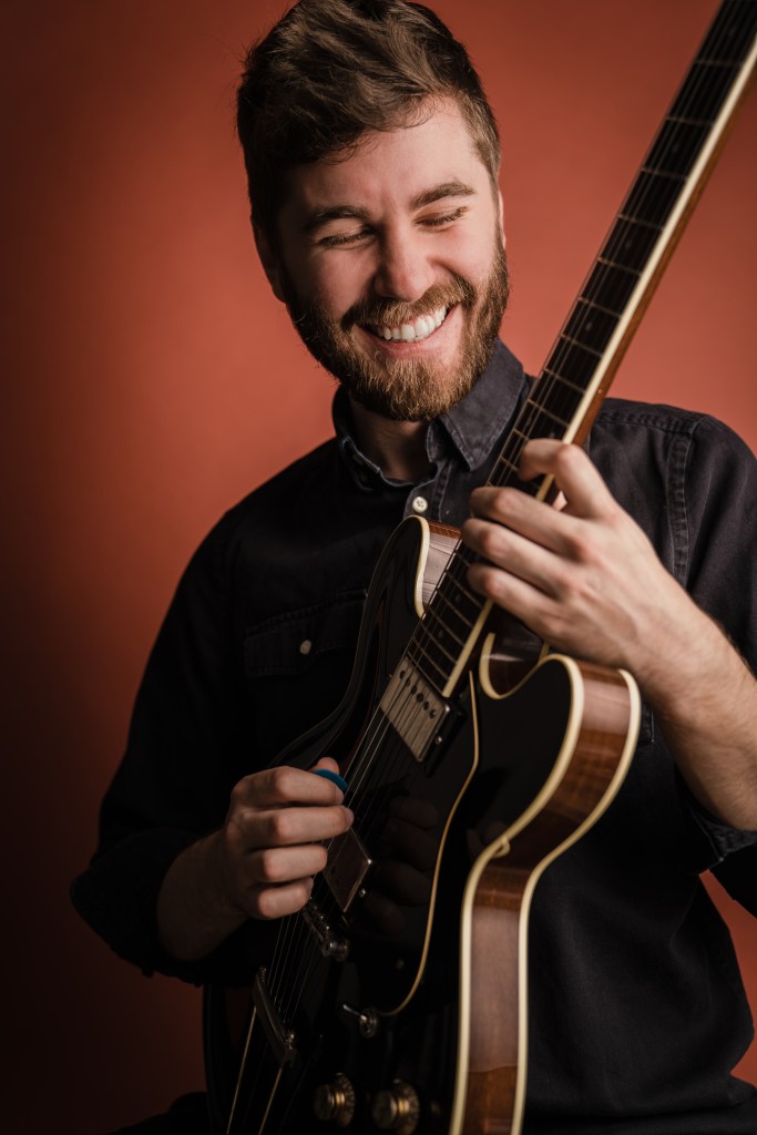 Max Light playing guitar with red background looking happy