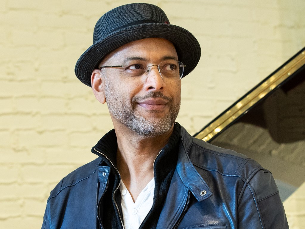 A mid-shot on Andy Milne, who looks off to the side, smiling. He wears a black jacket and stands against a cream-coloured brick background.