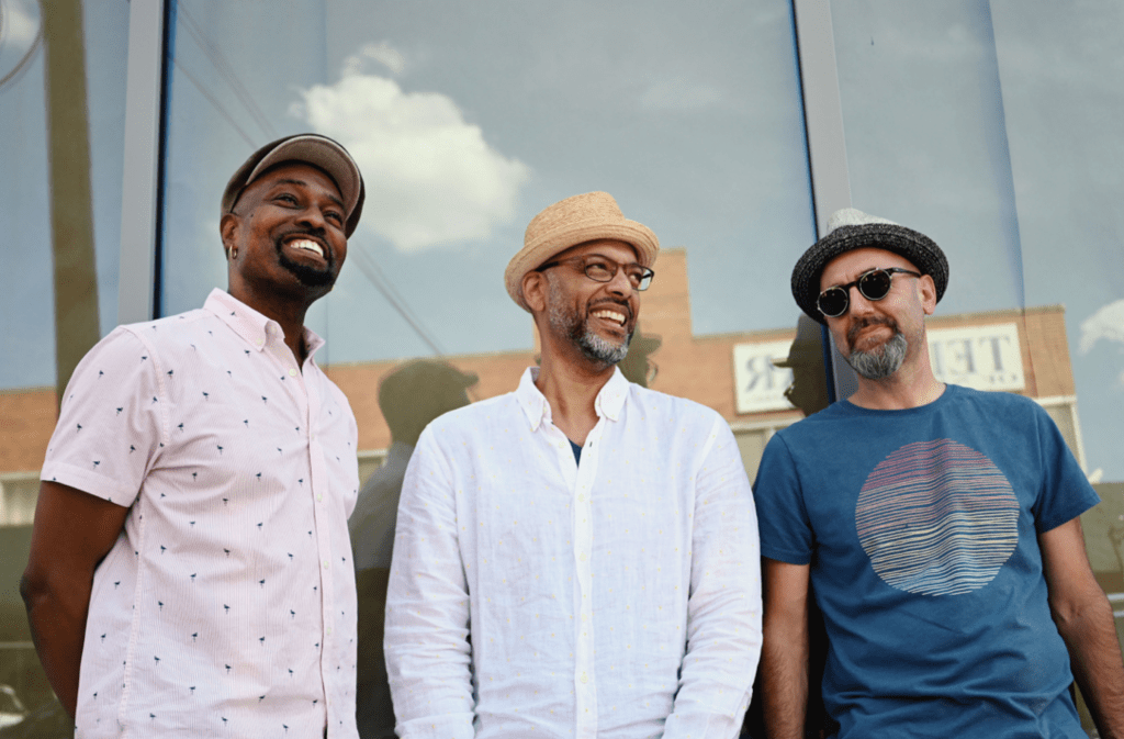 Clarence Penn, Andy Milne, and John Hébert stand in a line outside, against a reflective background which shows buildings and sky behind them. They are all smiling. 