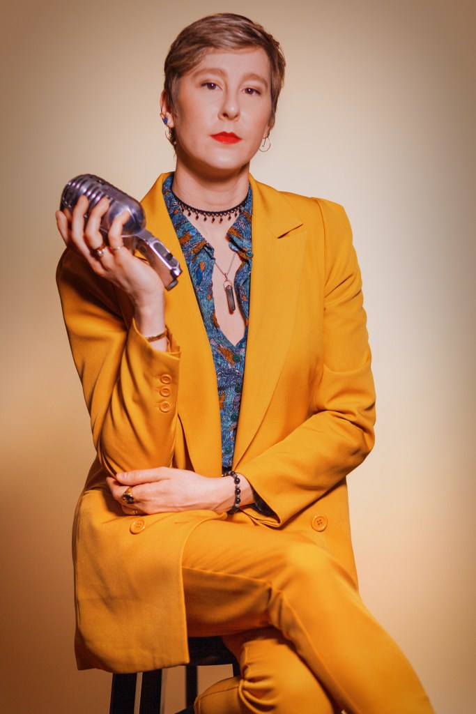Kelly green sits with her legs crossed, looking directly into camera wearing a mustard yellow suit. She holds an old microphone in one hand.