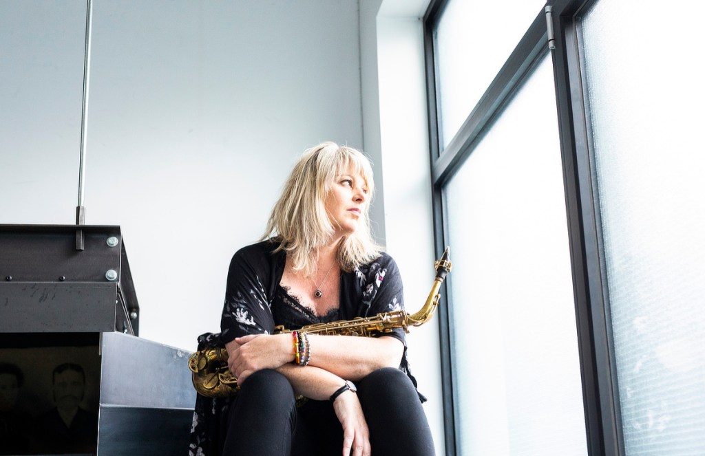 Christine Jensen sits holding her saxophone and looking out of a window which natural light pours through.